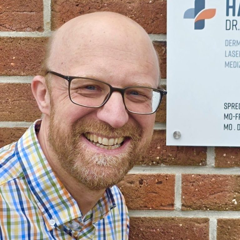 Mann mit Brille und kurzem Bart, trägt ein kariertes Hemd, lächelt vor moderner Architektur.